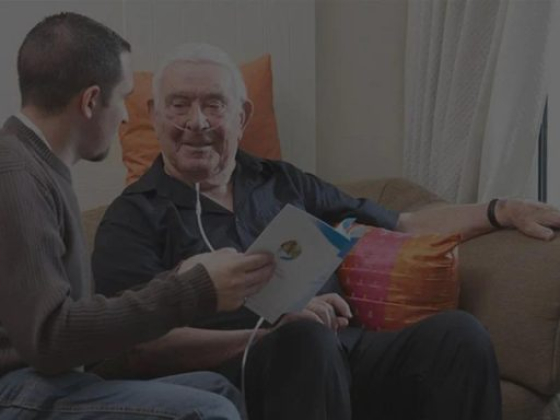Elderly man sits with caregiver, using oxygen concentrator